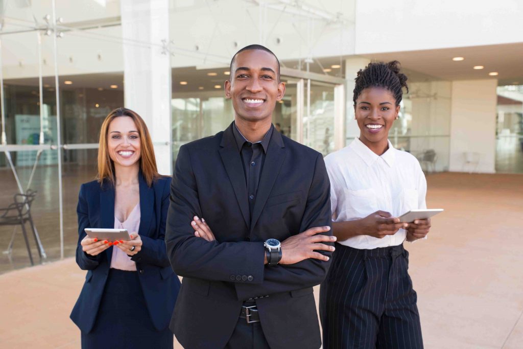 Happy cheerful business people posing in office hallway. Three multiethnic workers in formal suits standing for camera indoors and smiling. International cooperation concept